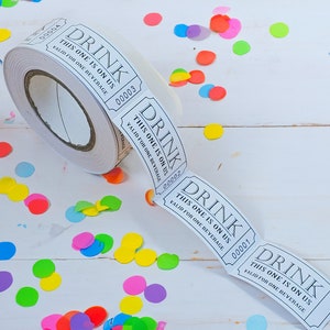 May include: A roll of white drink tickets with the text "DRINK THIS ONE IS ON US VALID FOR ONE BEVERAGE" printed in black. Multicoloured confetti is scattered around the roll on a white wooden surface.