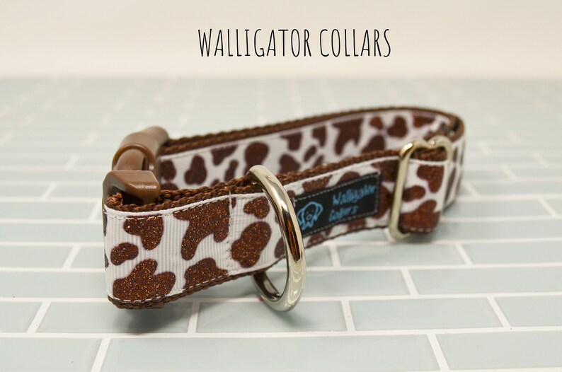 May include: A brown and white dog collar with a cow print pattern and glitter accents. The collar has a brown plastic buckle and a silver metal D-ring. The collar is labeled "Walligator Collars".