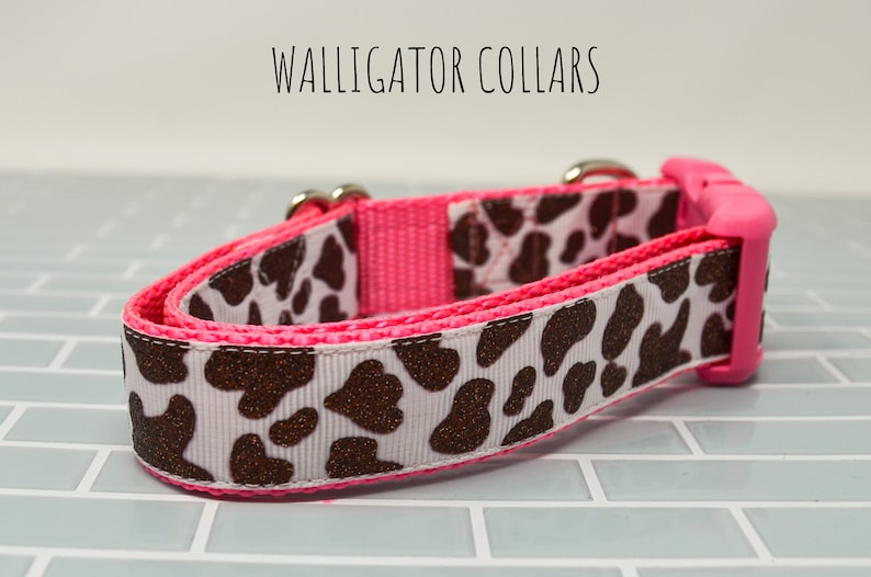 May include: A pink and white dog collar with a brown cow print pattern. The collar has a pink buckle and silver hardware. The text "WALLIGATOR COLLARS" is visible at the top of the image.