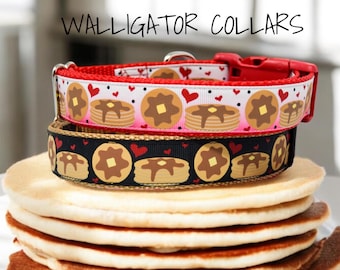 May include: Two dog collars with a pancake and heart pattern. The top collar is red with white and pink accents. The bottom collar is black with gold accents. The collars are stacked on top of a pile of pancakes.
