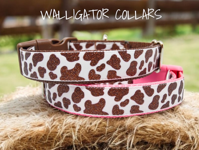 May include: Two dog collars with a brown and white cow print pattern. The top collar is brown with a pink accent. The bottom collar is pink with a brown accent. The collars are made of fabric with a glitter finish. The text "WALLIGATOR COLLARS" is visible at the top of the image.