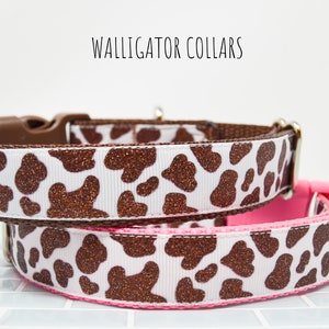 May include: Two dog collars with a brown and white cow print pattern. The collars are made of a white fabric with brown glitter spots. The top collar has a brown buckle and the bottom collar has a pink buckle.