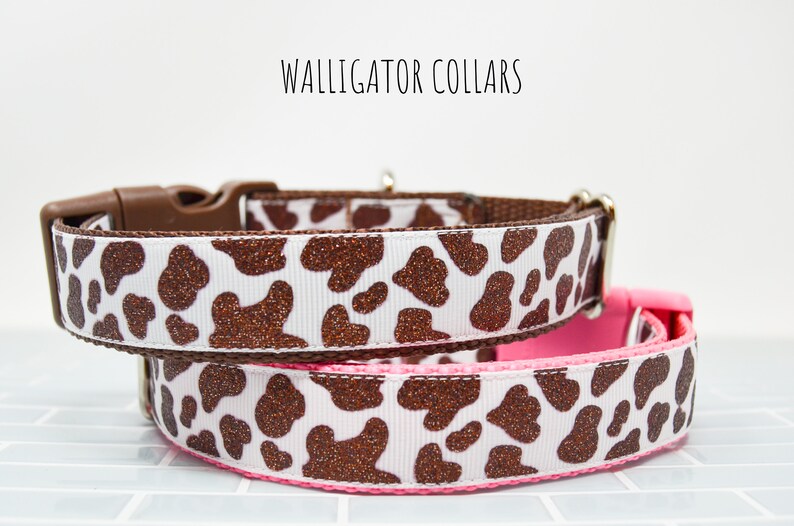 May include: Two dog collars with a brown and white cow print pattern. The collars are made of a white fabric with brown glitter spots. The top collar has a brown buckle and the bottom collar has a pink buckle.