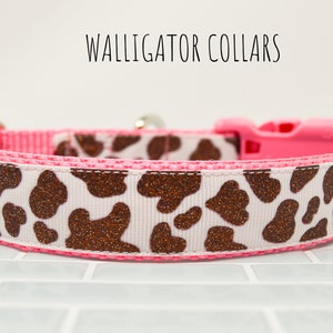 May include: A pink and white dog collar with a brown glitter cow print pattern. The collar has a pink plastic buckle and a silver D-ring. The text "WALLIGATOR COLLARS" is visible above the collar.