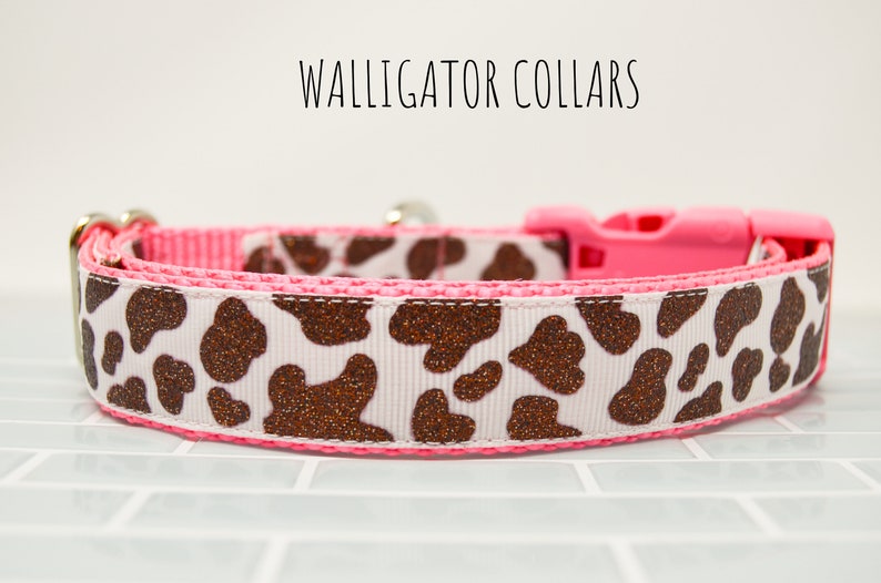 May include: A pink and white dog collar with a brown glitter cow print pattern. The collar has a pink plastic buckle and a silver D-ring. The text "WALLIGATOR COLLARS" is visible above the collar.