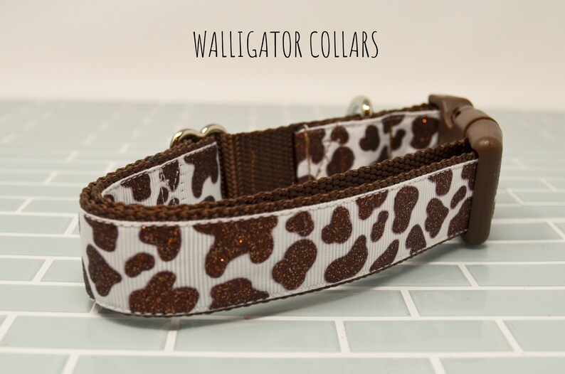 May include: A brown and white dog collar with a cow print pattern and brown glitter accents. The collar has a silver buckle and a brown plastic slider.