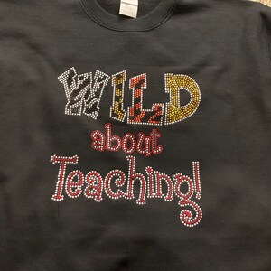 May include: Black sweatshirt with the phrase "WILD about Teaching!" in rhinestone lettering. The word "WILD" has animal print patterns, and the rest of the text is in red and white rhinestones. A crew neck sweatshirt.