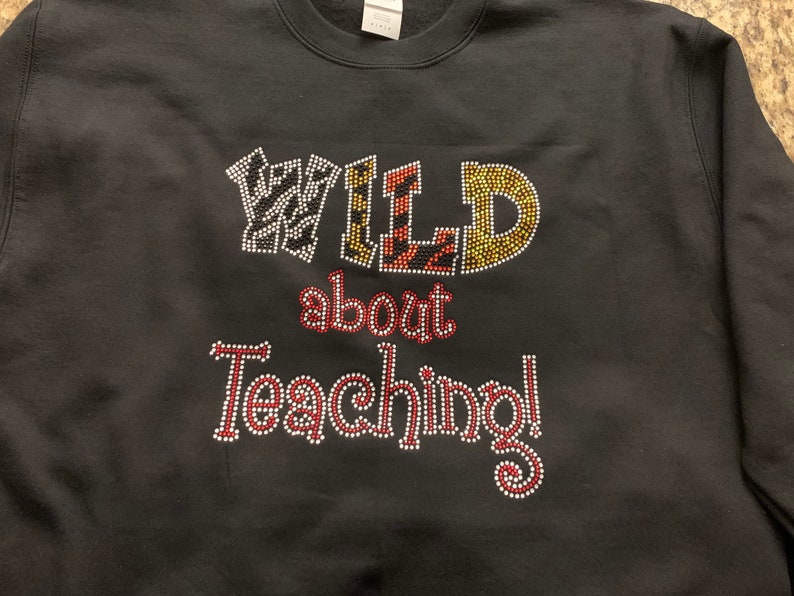 May include: Black sweatshirt with the phrase "WILD about Teaching!" in rhinestone lettering. The word "WILD" has animal print patterns, and the rest of the text is in red and white rhinestones. A crew neck sweatshirt.