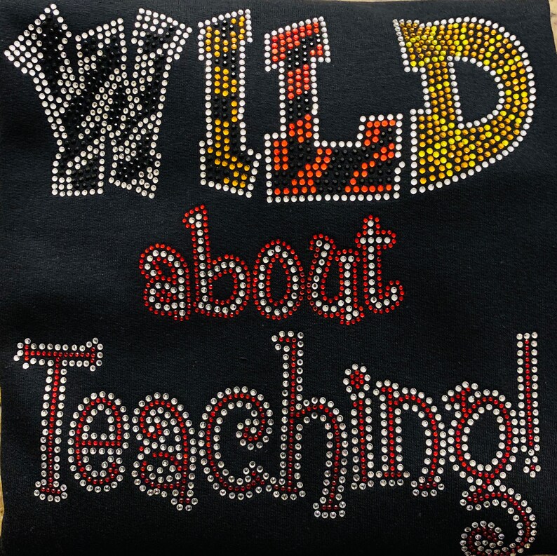 May include: Black sweatshirt with the phrase "WILD about Teaching" in rhinestone lettering. The word "WILD" has a leopard print design. The words are outlined in red and white rhinestones.