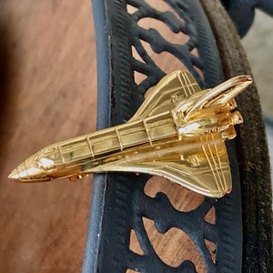 Puede incluir: Un pin de metal de color dorado con forma de transbordador espacial. El pin está detallado con líneas y un pequeño motor en la parte posterior.