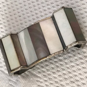 Puede incluir: Un anillo de servilleta de plata y nácar con un diseño ondulado. El anillo está hecho de varias piezas de nácar en diferentes tonos de blanco, gris y negro.