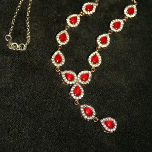 Puede incluir: Un collar de cadena de plata con piedras rojas en forma de lágrima engastadas en plata. Las piedras están rodeadas de piedras más pequeñas y transparentes.