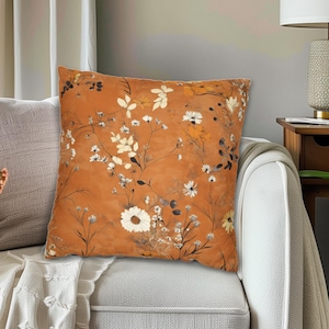 May include: A square decorative pillow with a floral pattern in shades of orange, white, and brown. The pillow is placed on a white sofa, with a neutral-colored pillow behind it. The overall theme is home decor.