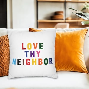Puede incluir: Cojín blanco con el texto "LOVE THY NEIGHBOR" en letras de colores. El cojín está sobre un sofá blanco, junto a un cojín naranja y un cojín marrón texturizado. Una planta está en el fondo.