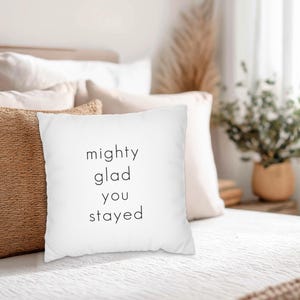 May include: White square pillow with the text "mighty glad you stayed" in a black, minimalist font. The pillow is on a bed with other pillows in shades of white and brown. The bed has a white comforter and a woven brown blanket.