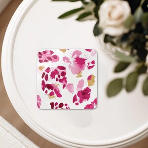 May include: Square coaster with a white background and a pattern of pink and gold abstract shapes. The coaster is sitting on a white table, with a vase of flowers in the background.