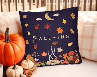May include: A navy blue throw pillow with a whimsical design featuring a crescent moon, clouds, and falling autumn leaves. The text "FALL-ING" is written in a stylized font in gold.