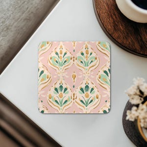 Puede incluir: Posavasos cuadrado con un patrón floral repetido en verde, dorado y crema sobre un fondo rosa. El posavasos está sobre una superficie blanca, con una taza de café y elementos decorativos en el fondo.