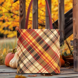 May include: A plaid tote bag with burgundy straps. The bag features a diagonal pattern in shades of red, orange, yellow, and brown. The background includes pumpkins and fall foliage.