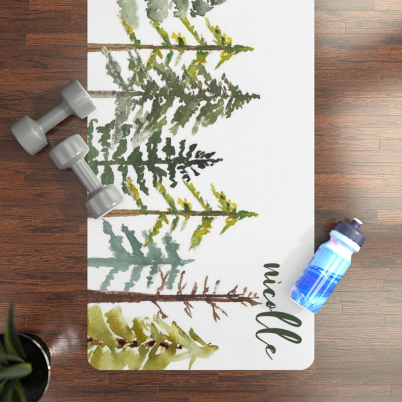 May include: A white yoga mat with a watercolor design of green pine trees. The name "nicole" is written in black cursive at the bottom of the mat.