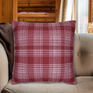 May include: A square decorative pillow with a red and white plaid pattern. The pillow is placed on a beige sofa, with a brown blanket visible on the left side. The plaid design features intersecting white lines on a red background.
