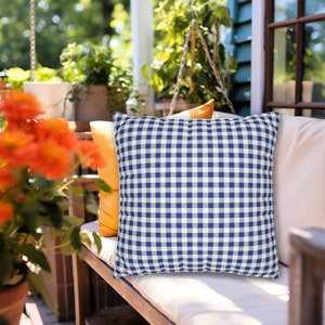 Puede incluir: Un coj&iacute;n de tiro de gingham azul y blanco en un banco de exterior blanco. El coj&iacute;n est&aacute; hecho de una tela suave y c&oacute;moda y tiene una forma cuadrada.