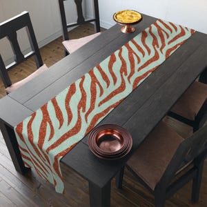 May include: A rectangular table runner with a zebra stripe pattern in shades of red and light blue. The runner is placed on a dark wooden table, with a stack of copper-colored plates and a pie on a stand.