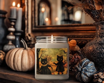 Może przedstawiać: Zapalone świeca w szklanym słoiku z etykietą o tematyce Halloween, przedstawiającą dwie czarne postacie kota. Świeca stoi na drewnianej powierzchni z dyniami, świecami i elementami dekoracyjnymi.