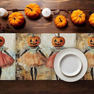 May include: A Halloween-themed table setting with orange pumpkins, white candles, and a decorative runner. The runner features a repeating pattern of figures with pumpkin heads and ballerina costumes. A white plate with a gold rim is also present.