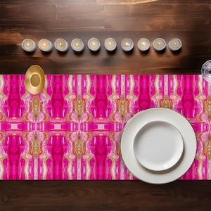 Pode incluir: Uma mesa com um caminho de mesa com padrão rosa e dourado vibrante. Um prato branco com borda dourada está sobre o caminho de mesa, junto com um carregador dourado e várias velas acesas. A mesa é de madeira escura.