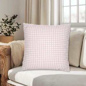 May include: A square decorative pillow with a pink and white gingham pattern. The pillow is placed on a white couch with other neutral-colored pillows. A wicker chair and a houseplant are visible in the background.