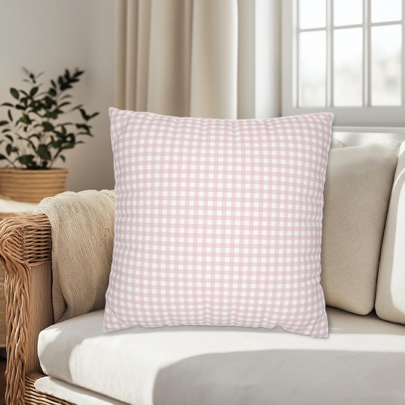 May include: A square decorative pillow with a pink and white gingham pattern. The pillow is placed on a white couch with other neutral-colored pillows. A wicker chair and a houseplant are visible in the background.