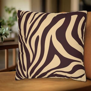 Op de afbeelding: Vierkant kussen met een zebra streep patroon. Het kussen heeft een crèmekleurige achtergrond met donkerbruine strepen, wat een gedurfd, door dieren geïnspireerd ontwerp creëert. Het kussen ligt op een bruin oppervlak.