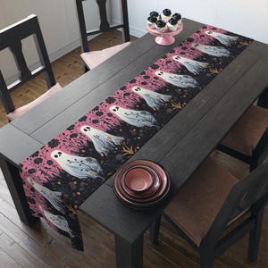 May include: A Halloween-themed table runner featuring a repeating pattern of white ghost figures with floral accents on a black and pink background. The runner is displayed on a dark wooden table, with matching plates and cupcakes.