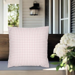 May include: A square decorative pillow with a pink and white gingham pattern. The pillow is placed on a wooden bench, suggesting outdoor or patio use. The pillow's design is a classic, country-style pattern.
