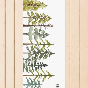 May include: A white yoga mat with a watercolor design of a forest of green trees. The name "Nicole" is written in green at the bottom right corner of the mat.