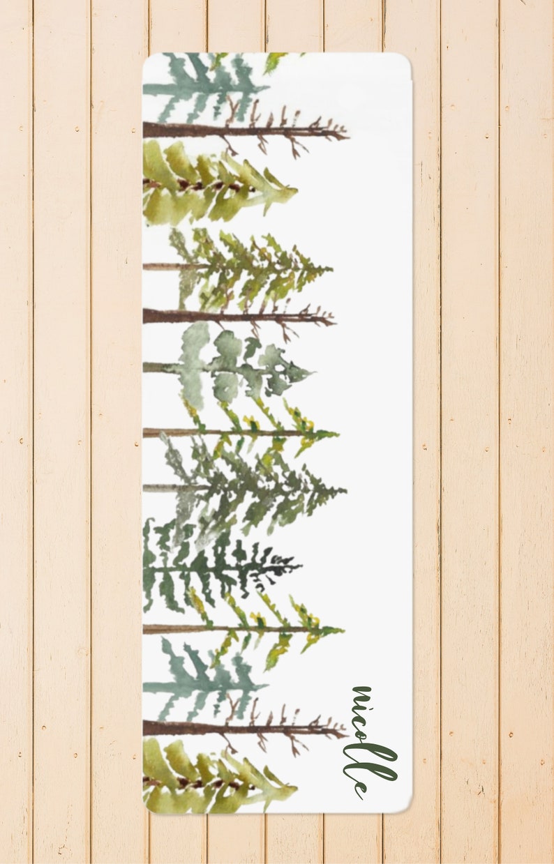 May include: A white yoga mat with a watercolor design of a forest of green trees. The name "Nicole" is written in green at the bottom right corner of the mat.