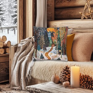 May include: A throw pillow featuring a colourful illustration of a snowy woodland scene with a small cottage. The pillow is on a sofa with a blanket and a candle.