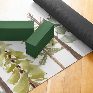May include: A white yoga mat with a watercolor design of green trees. Two green yoga blocks and a rolled up black yoga mat are on top of the mat.