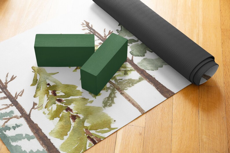 May include: A white yoga mat with a watercolor design of green trees. Two green yoga blocks and a rolled up black yoga mat are on top of the mat.