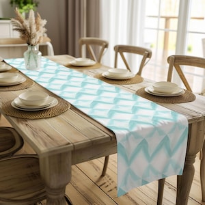 May include: A rectangular table runner with a white background and a repeating geometric pattern in shades of light blue. The runner is placed on a wooden table set for a meal, with plates, placemats, and chairs visible.