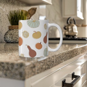 May include: White ceramic mug with a fall-themed design featuring gourds, pumpkins, and ladybugs in shades of orange, yellow, green, and brown.