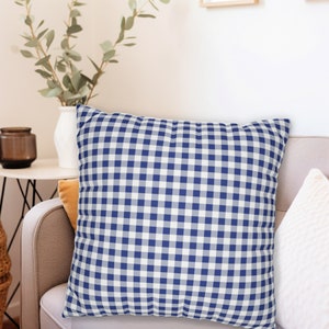 Puede incluir: Una almohada de tiro de gingham azul y blanco con un patr&oacute;n cuadrado. La almohada est&aacute; sentada en un sof&aacute; marr&oacute;n claro.