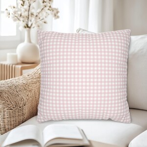 May include: A square decorative pillow with a pink and white gingham pattern. The pillow is on a white sofa, with a wicker chair and a vase of white flowers in the background. The pillow is a soft, home decor item.