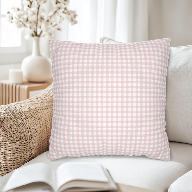 May include: A square decorative pillow with a pink and white gingham pattern. The pillow is on a white sofa, with a wicker chair and a vase of white flowers in the background. The pillow is a soft, home decor item.