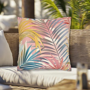 May include: Square decorative pillow with a tropical palm leaf pattern. The pillow features a vibrant design with yellow, pink, blue, and gray leaves on a white background. The pillow is placed on a wicker outdoor sofa.