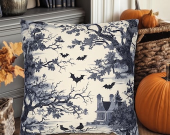 May include: Square decorative pillow with a Halloween-themed toile design. The pillow features a cream background with a dark blue illustration of a haunted house, bats, trees, and birds. The pillow is placed on a wooden surface.