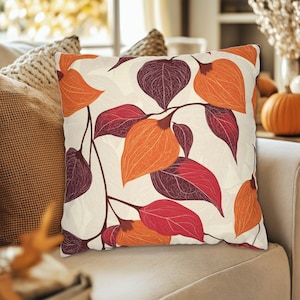 Pode incluir: Almofada decorativa quadrada com fundo creme e um padrão repetido de folhas e vagens de sementes laranja e bordô. A almofada está em um sofá bege, sugerindo um tema de outono.