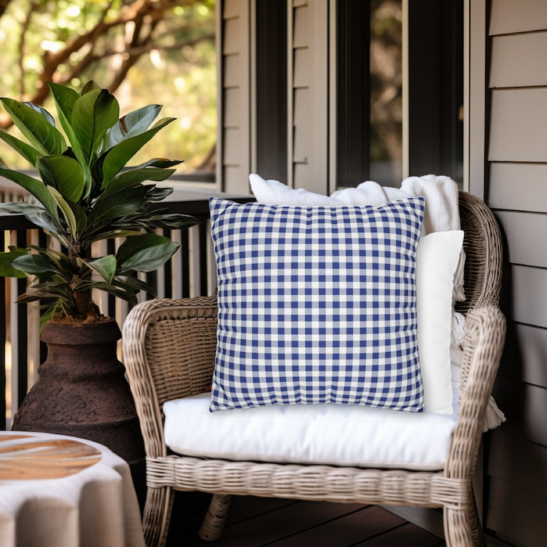 Puede incluir: Una silla de mimbre con un coj&iacute;n blanco y una almohada de cuadros azules y blancos. La silla est&aacute; en un porche con suelo de madera y una planta en maceta.