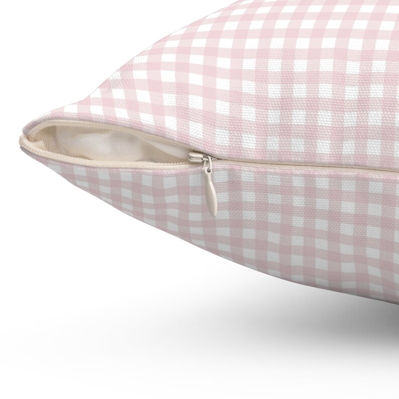 May include: A close-up of a square pillow with a pink and white gingham pattern. The pillow has a zipper closure on one side. The fabric is a soft, woven material. The pillow is a decorative home accessory.